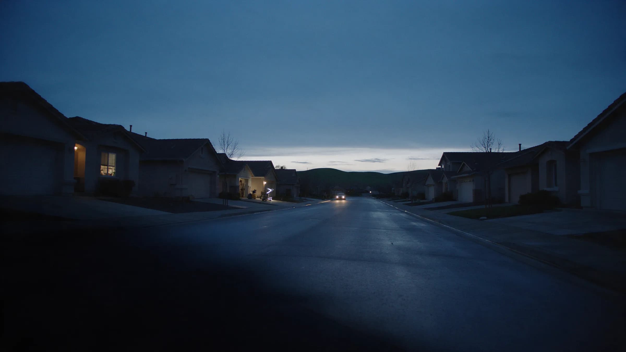 A quiet Gilroy street about twenty minutes before sunrise, one kitchen window lit, a single car coming up the road with its headlights on and nobody in the frame.